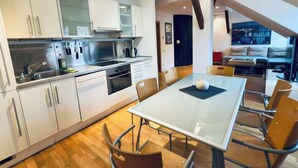 Une cuisine moderne avec des armoires blanches, une hotte de cuisinière en acier inoxydable et un coin repas doté d’une table en verre et de chaises en métal.