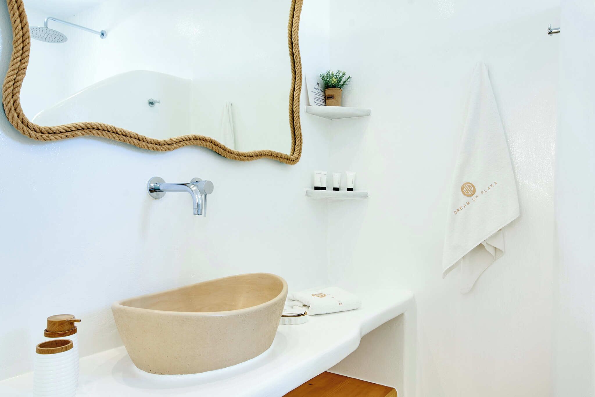 A bathroom with a large beige sink, a mirror with a rope frame, a shower, and a small shelf with toiletries.