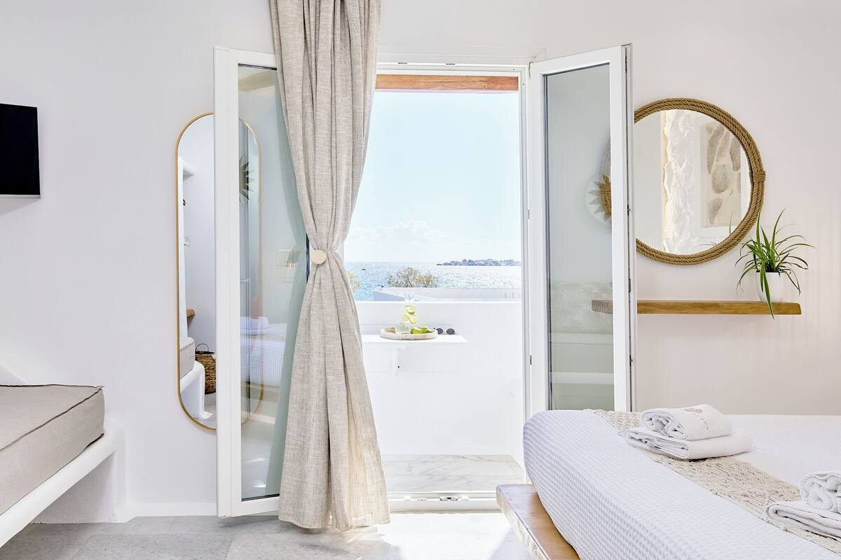 A hotel room with a bed, a bedside table, a mirror, and a view of the sea through an open door.