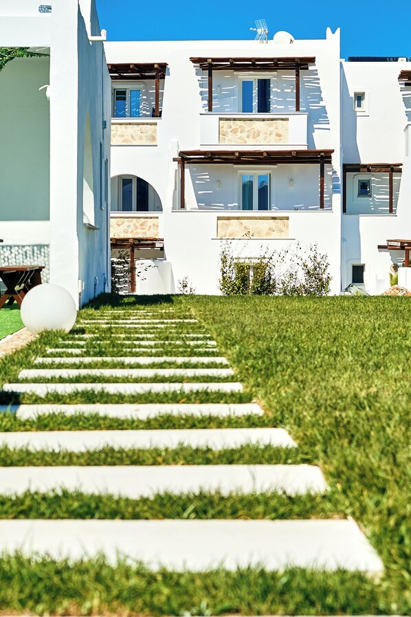 A modern building with a white facade, multiple balconies, and a well-maintained lawn with a stone pathway leading to the entrance. - Dream on Plaka (Naxos)
