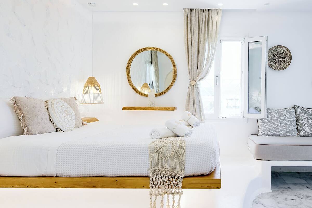 A modern bedroom with a large bed, a bedside lamp, a mirror, and a view of the outdoors.