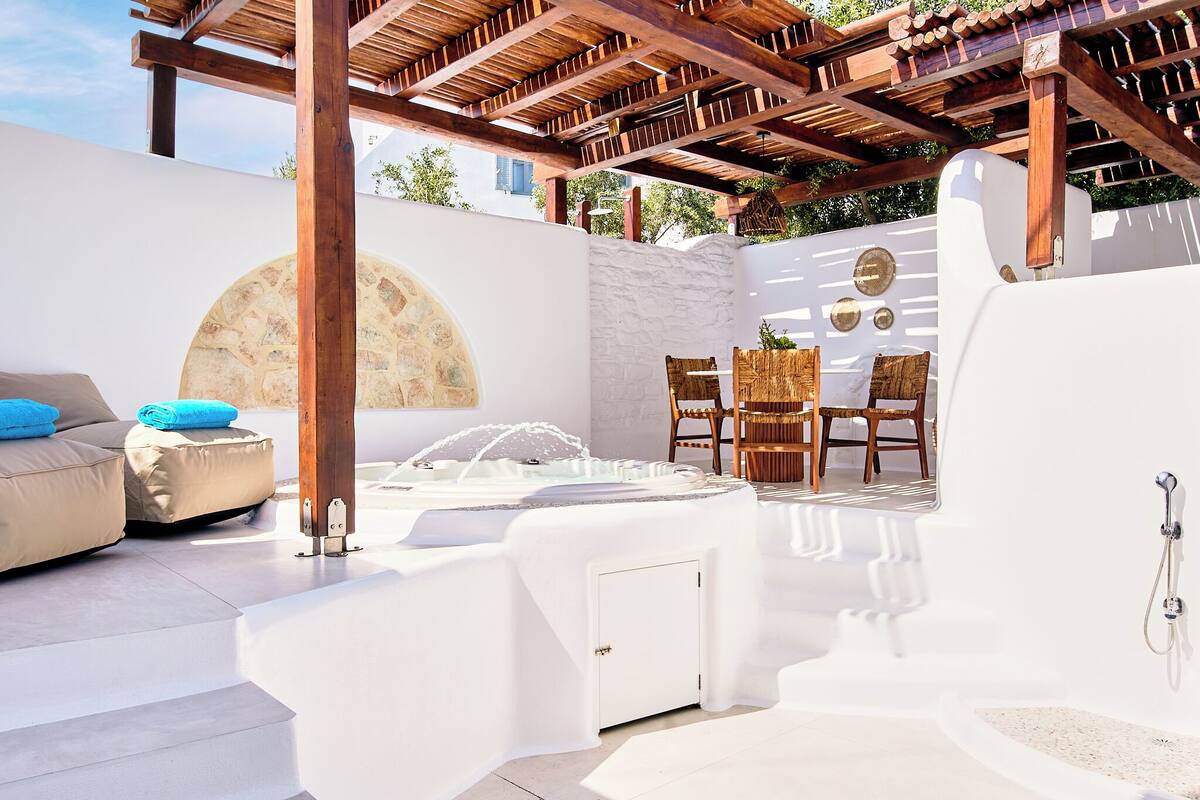 A sunlit outdoor patio with a stone wall, wooden pergola, a white outdoor seating area, and a hot tub.