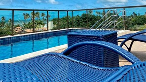 A poolside area with blue wicker lounge chairs, a clear blue sky, and a view of palm trees and the ocean. - Green Porto 06 (Ipojuca)