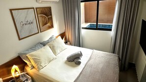 A hotel room with a bed, a nightstand with a lamp, a wall-mounted air conditioner, and a window with curtains. - Green Porto 06 (Ipojuca)