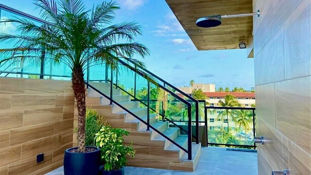 A modern staircase with glass railings leading to a balcony with a view of palm trees and buildings. - Green Porto 06 (Ipojuca)