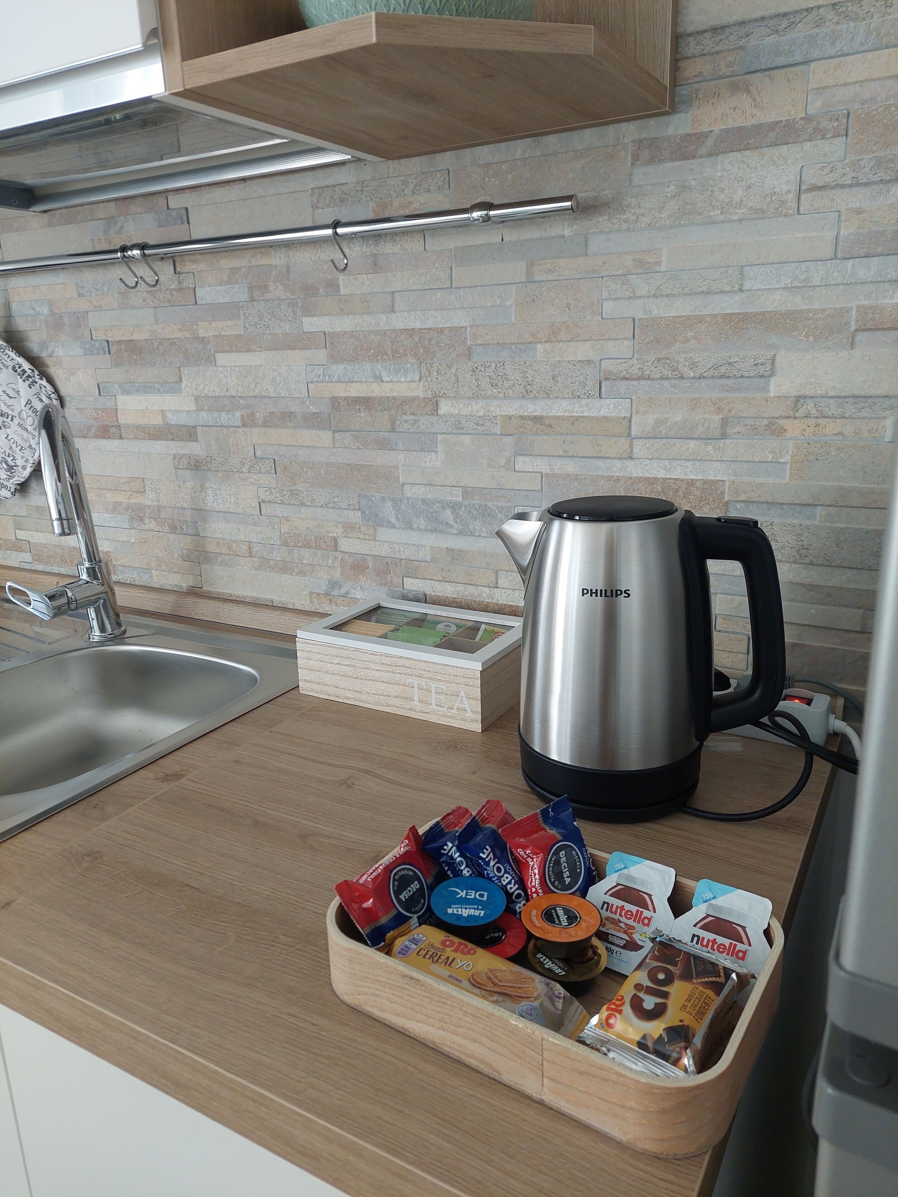 A kitchen with a stainless steel kettle, a wooden tray with Nutella and cereal bars, and a box labeled "TEA".