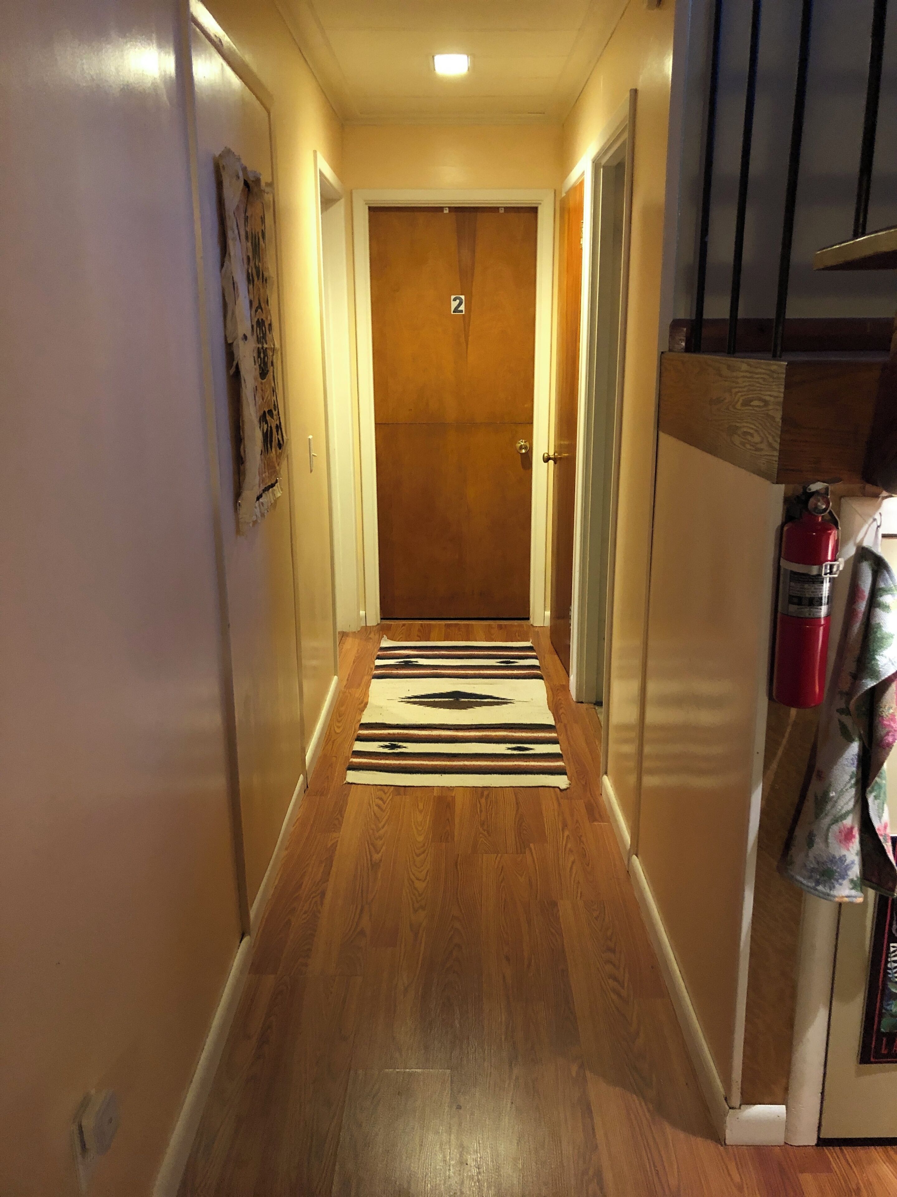 A narrow hallway with wooden flooring, a door with the number 2, and a fire extinguisher on the right.