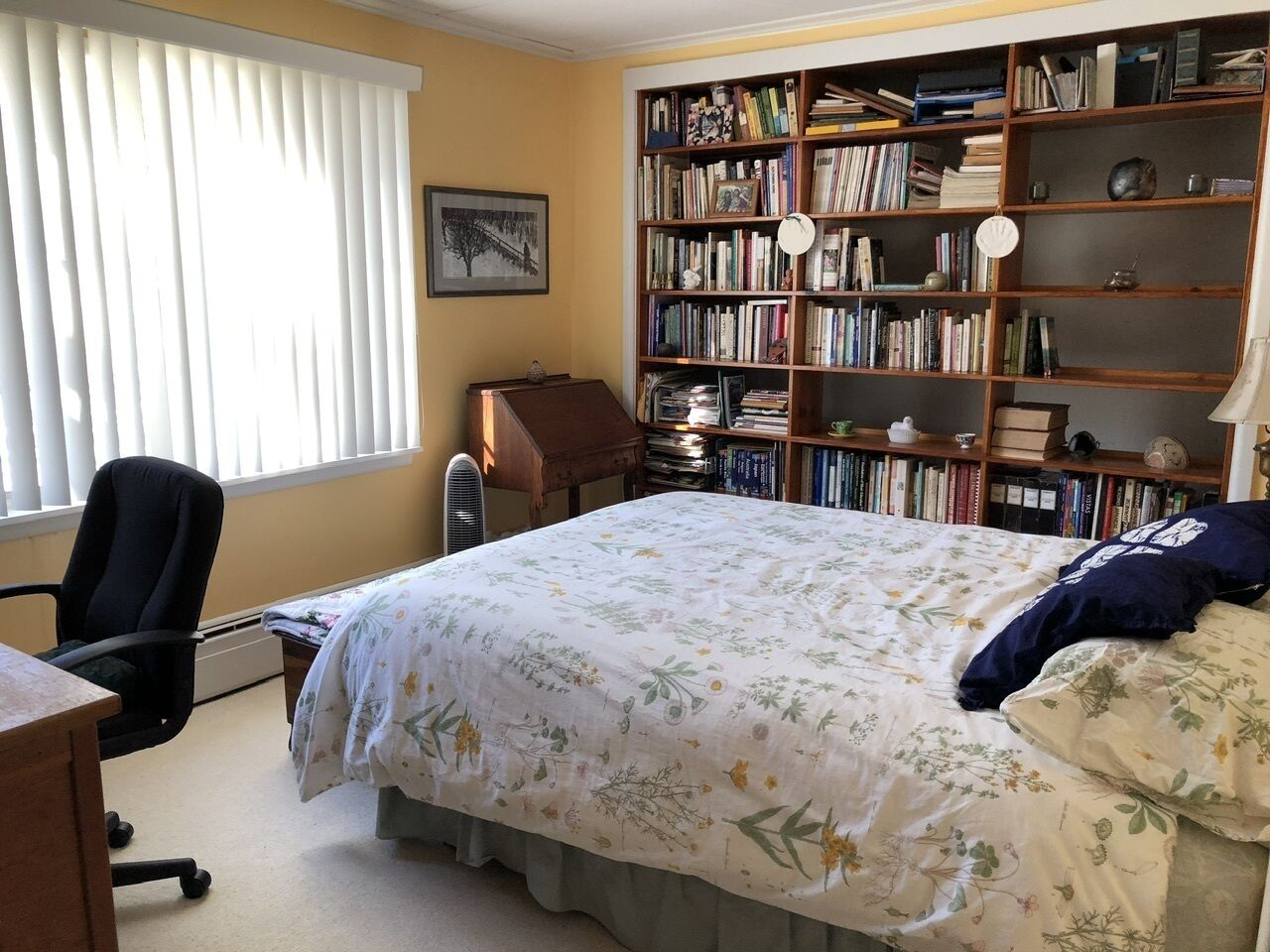 Une chambre à coucher comprenant un lit, un bureau, une chaise, une bibliothèque et une fenêtre avec des stores.
