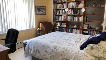 Une chambre à coucher comprenant un lit, un bureau, une chaise, une bibliothèque et une fenêtre avec des stores.