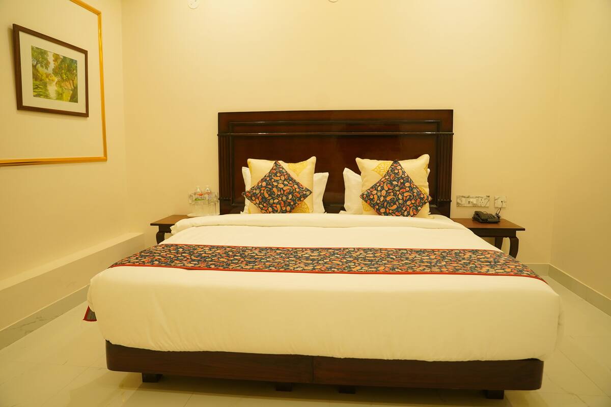 A neatly made bed with a wooden headboard, white bedding, and decorative pillows. There is a small bedside table with a lamp and a framed picture on the wall.