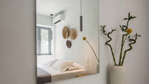 A modern bedroom with a large mirror, a bed with white linens, a decorative wall hanging, and a vase with a plant. - Rivière Residence Paros   (Paros)