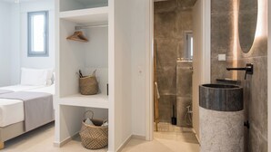 A modern hotel room with a bed, a built-in wardrobe, a bathroom with a large sink and mirror, and a shower area. - Rivière Residence Paros   (Paros)