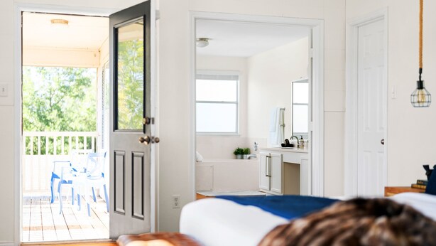 Quarto com chão de madeira, uma cama, banheiro com pia e espelho, e varanda com cadeiras.