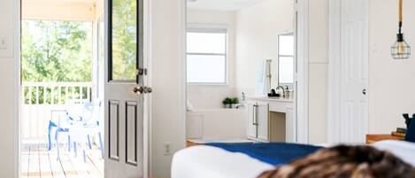 Une chambre avec un plancher en bois, un lit, une salle de bain avec un lavabo et un miroir, et un balcon avec des chaises.