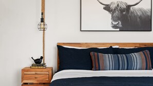 Quarto com cabeceira de madeira, travesseiro listrado, quadro de um touro, luminária de cabeceira e mesa de cabeceira de madeira com livros.