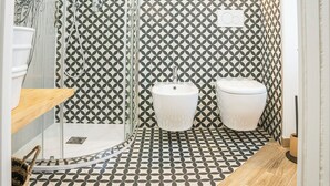 A bathroom with a geometric patterned tile floor and walls, a glass shower enclosure, a wooden shelf, and a white sink and toilet.
