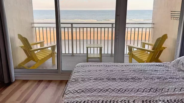 A balcony with a view of the beach, two yellow chairs, a small table, and a bed with patterned bedding. - The Lidiya Room, right on the famous Daytona Beach. (Daytona Beach)