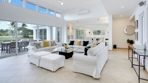 A spacious living area with a white sofa set, a coffee table, and a dining table with chairs. Large windows provide natural light and a view of the outdoors. - Two Luxury Villas in Cocotal for Large Groups – With Jacuzzi, Pools, Gym (Punta Cana)
