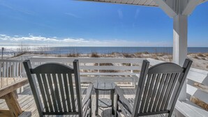 Un espace extérieur abrité, aménagé avec deux chaises en bois et une petite table, offrant une vue sur une plage avec des dunes de sable et l’océan.