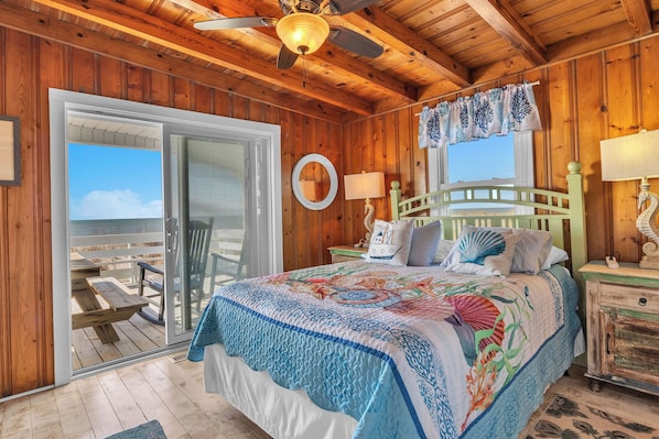 A wooden cabin bedroom with a bed, a nightstand, a window with curtains, a ceiling fan, and a view of the beach through a sliding glass door.
