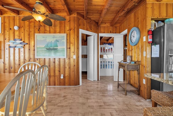 A wooden cabin interior with a dining table, chairs, a ceiling fan, a clock, and a refrigerator. - The Mermaid Cottage: Charming, Dog-Friendly and Oceanfront (Oak Island)