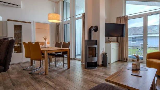 A modern living room with a dining area, a fireplace, a flat-screen TV, and large windows. - Ferienhaus mit Sauna im Ferienpark Mirow (Mirow)