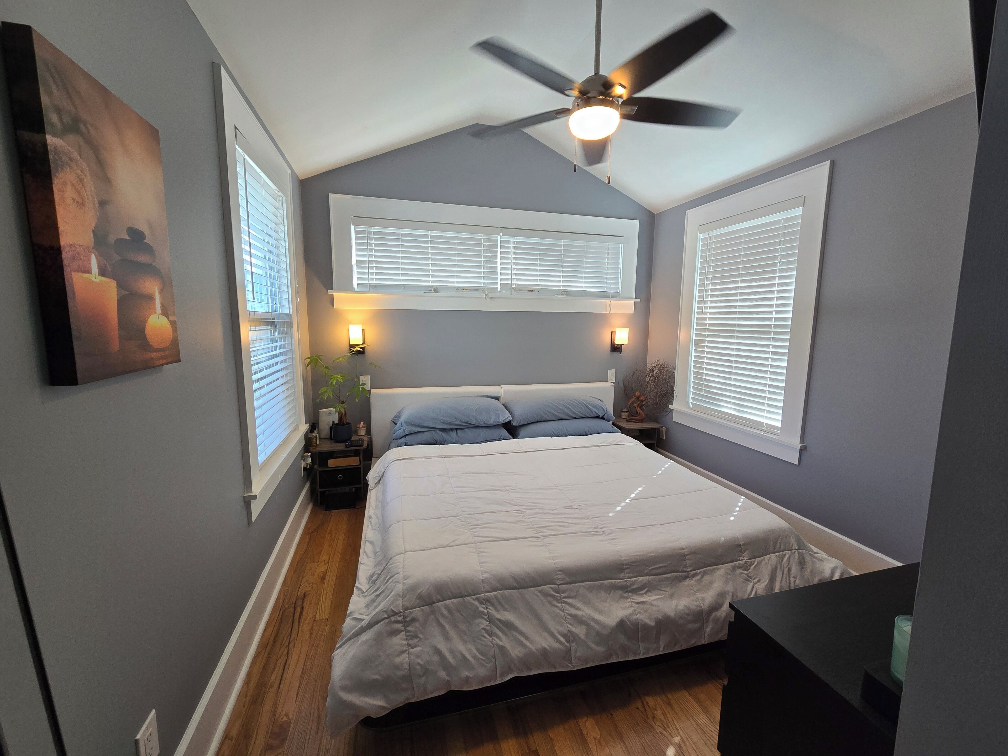 Ein Schlafzimmer mit Bett, Deckenventilator, Nachttischlampen, Fenstern mit Jalousien und einem an der Wand befestigten Regal.