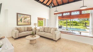 A spacious living room with two beige sofas, a coffee table, and a large window overlooking a pool and greenery.