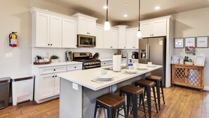 A modern kitchen with white cabinets, a central island with stools, stainless steel appliances, and pendant lighting. - Stunning Townhome in Zion Village with private hot tub and lazy river (Hurricane)
