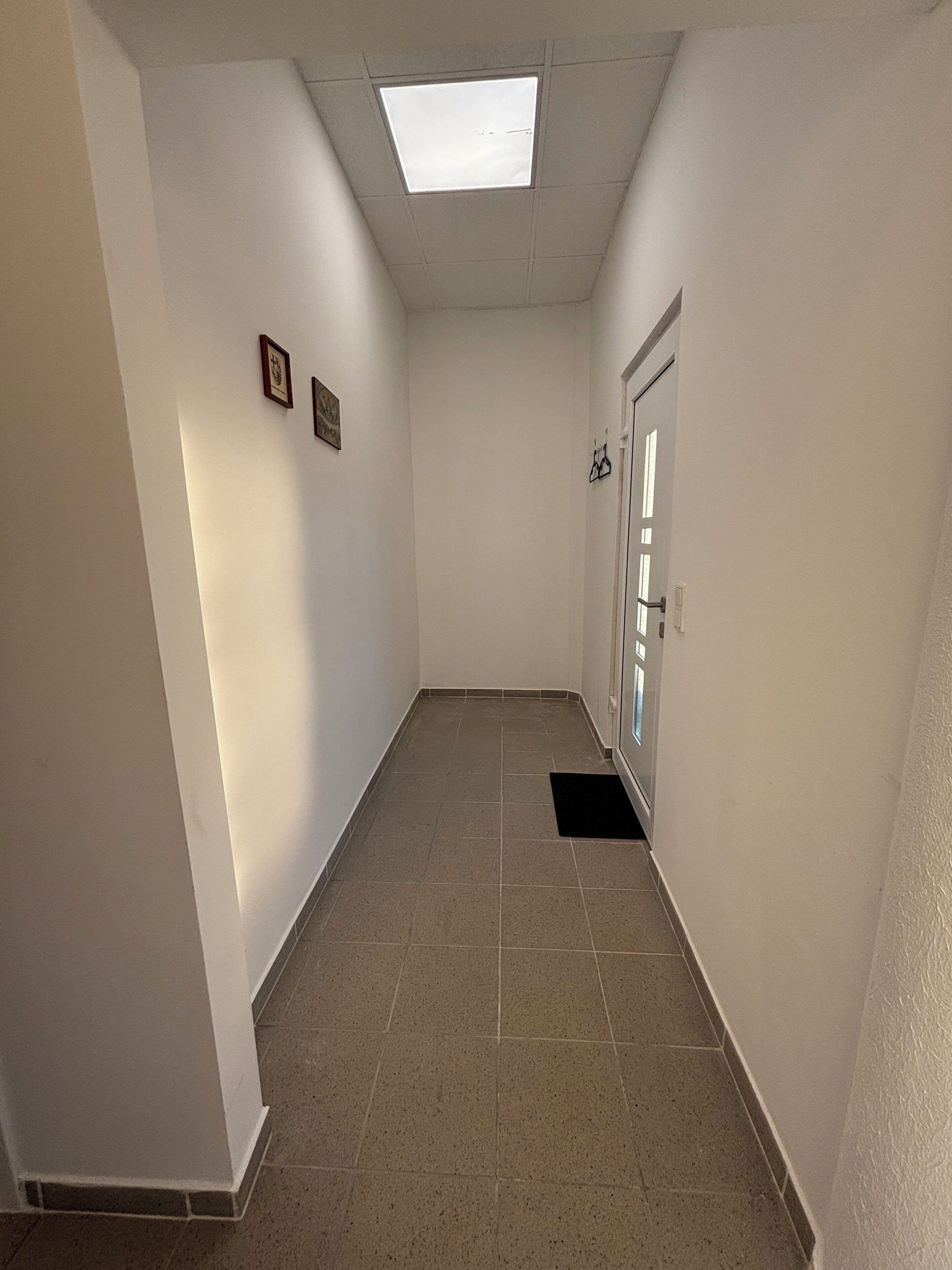 A narrow hallway with a tiled floor, white walls, a ceiling light, and a door with a window.
