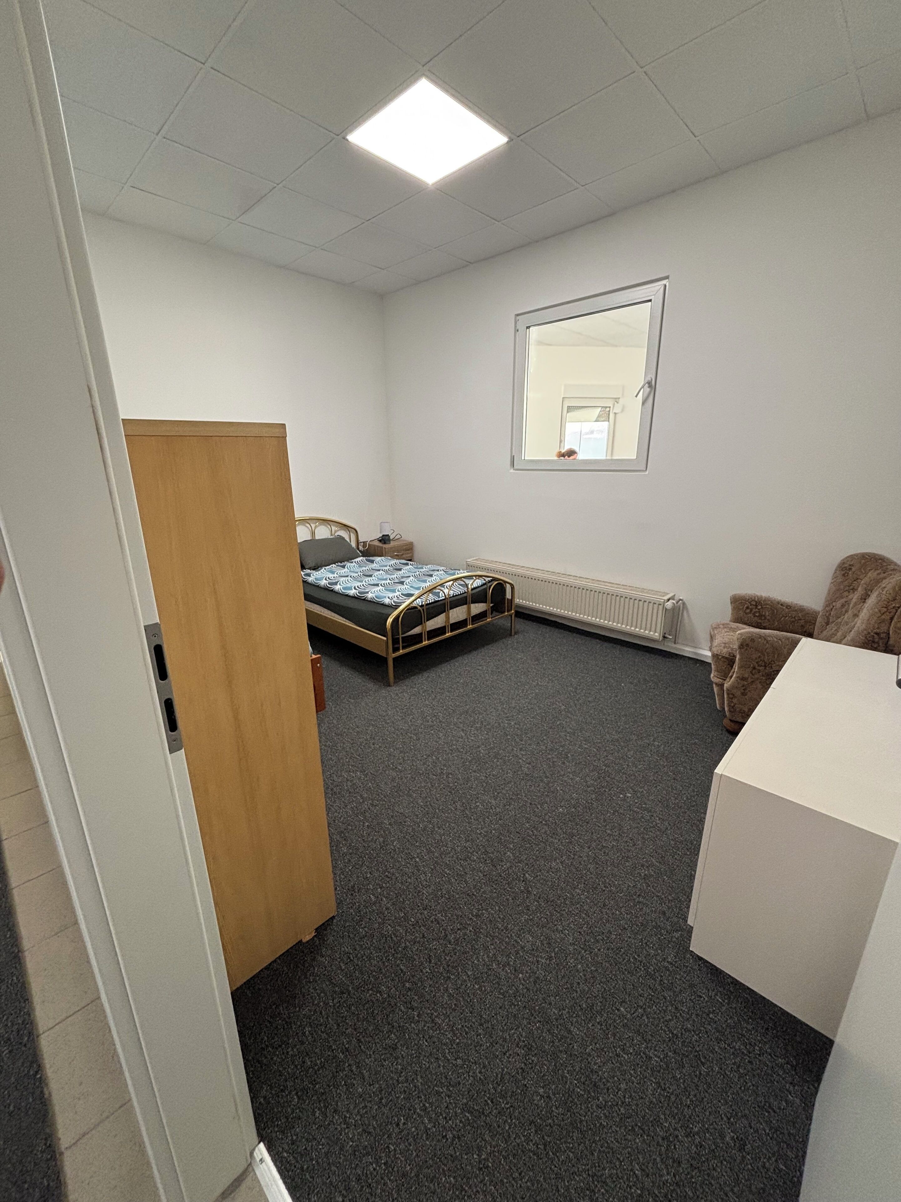 A small, sparsely furnished room with a bed, a desk, a radiator, and a window.