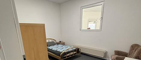 A small, sparsely furnished room with a bed, a desk, a radiator, and a window.