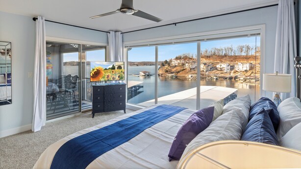 Een moderne slaapkamer met een groot bed, een plafondventilator, een televisie en uitzicht op een meer en gebouwen.