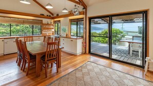 A spacious kitchen with wooden cabinets, a dining table, and chairs. There is a large sliding glass door leading to an outdoor deck with a view of the ocean. - Two Many Birds - Seal Rocks (Seal Rocks)