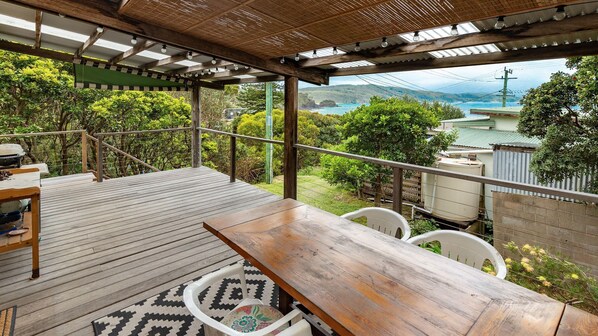 A wooden deck with a table and chairs, overlooking a scenic view of a coastal area with hills and a body of water. - Two Many Birds - Seal Rocks (Seal Rocks)