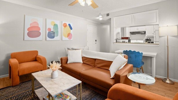 Una sala de estar con un sofá marrón, un sillón naranja y una mesa de centro. Se observa un ventilador de techo, una zona de cocina con armarios blancos y una lámpara de pie.