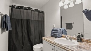 Un baño con cortina de ducha, lavamanos, espejo y una pila de toallas.