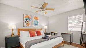Un dormitorio con cama, dos mesitas de noche, un escritorio, ventilador de techo y cuadros en la pared.