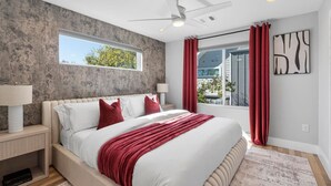 A modern bedroom with a large bed, a ceiling fan, a window with red curtains, and a view of the outdoors. - Spacious Austin Home Base| Sleeps 12+ | Near DT (Austin)