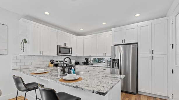 A modern kitchen with white cabinets, a stainless steel refrigerator, a granite countertop, and two black bar stools. - Spacious Austin Home Base| Sleeps 12+ | Near DT (Austin)