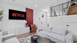 A modern living room with a large flat-screen TV displaying "NETFLIX," a white sofa, a brown leather chair, a black coffee table, and a staircase with a metal railing. - Spacious Austin Home Base| Sleeps 12+ | Near DT (Austin)