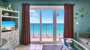A living room with a television, a glass table, and a sofa. There is a balcony with a view of the ocean.