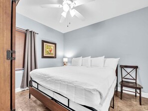 Ein Schlafzimmer mit Bett, Deckenventilator, Nachttisch mit Lampe, gerahmtes Bild an der Wand und Fenster mit Jalousien.