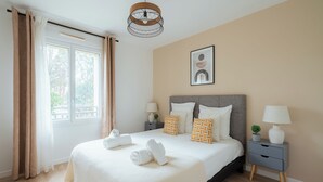 Ein ordentlich bezogenes Bett mit weißen Leinen und aufgerollten Handtüchern, ein Nachttisch mit einer Lampe und ein gerahmtes Bild an der Wand.
