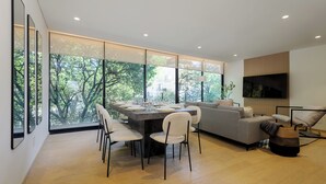 A modern living room with a dining area, large windows, a sofa, and a flat-screen TV.