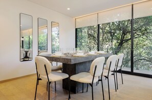 A modern dining area with a large table set for six, surrounded by white chairs. There are three large mirrors on the wall, and a large window offering a view of trees. - Saltillo 85 (Mexico City)