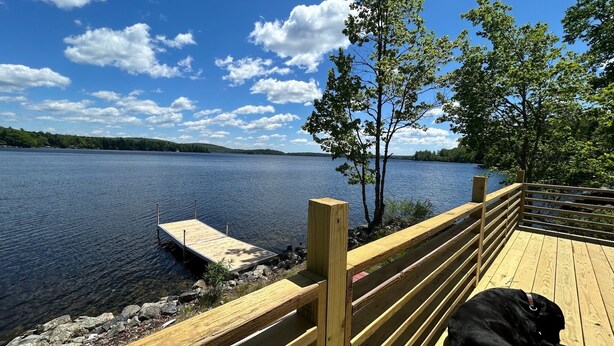 Una terraza de madera con un perro tumbado sobre ella, con vistas a un lago tranquilo y un muelle que se adentra en el agua.