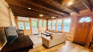 Interior de una cabaña de madera con sofá, televisor sobre una mesa y ventanas amplias.