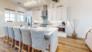 A modern kitchen with white cabinetry, a large island with bar stools, a chandelier, and a potted plant. - Gorgeous family beach home with beach & gulf views, balconies,  pools, & golf (Port Aransas)