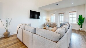 A modern living room with a sectional sofa, a flat-screen TV mounted on the wall, and a large window providing natural light. - Gorgeous family beach home with beach & gulf views, balconies,  pools, & golf (Port Aransas)